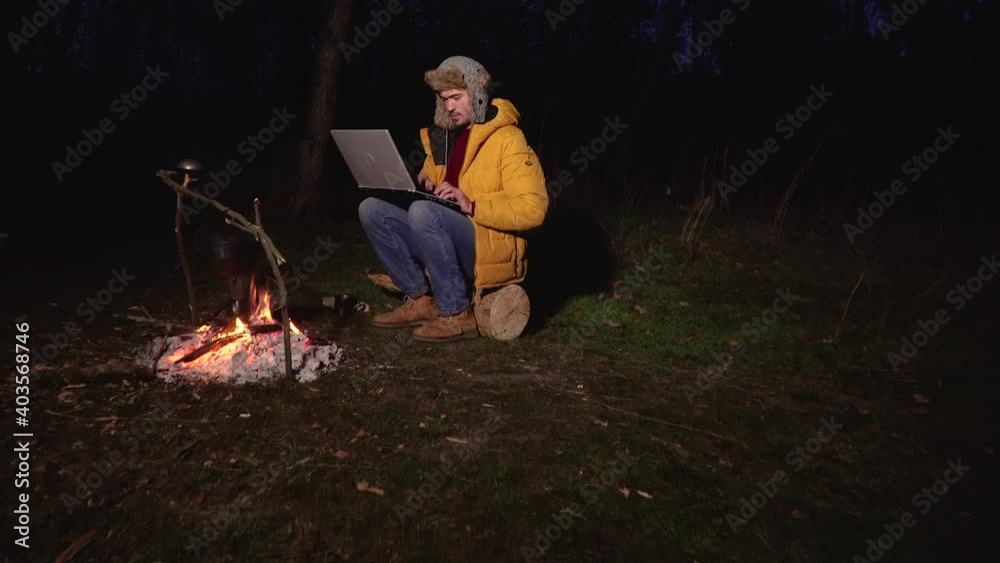 A young man works at a laptop near the fire in the forest in the evening. Freelance remote work concept. 4K A young man works at a laptop near the fire in the forest in the evening. Freelance remote work concept. 4K
