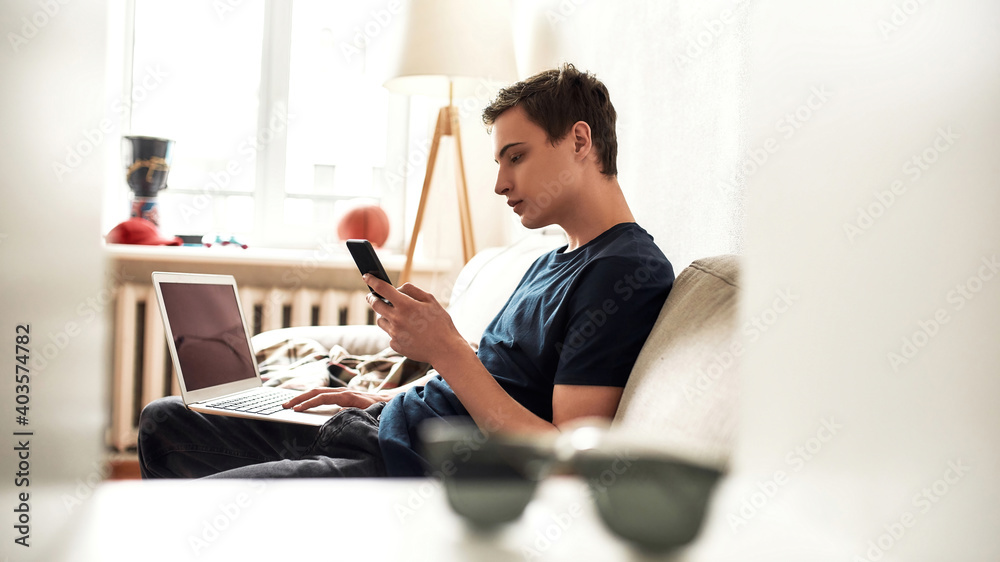 Young guy using laptop and mobile phone while spending time sitting on the couch at home. Technology, smartphone addiction concept Young guy using laptop and mobile phone while spending time sitting on the couch at home. Technology, smartphone addiction concept