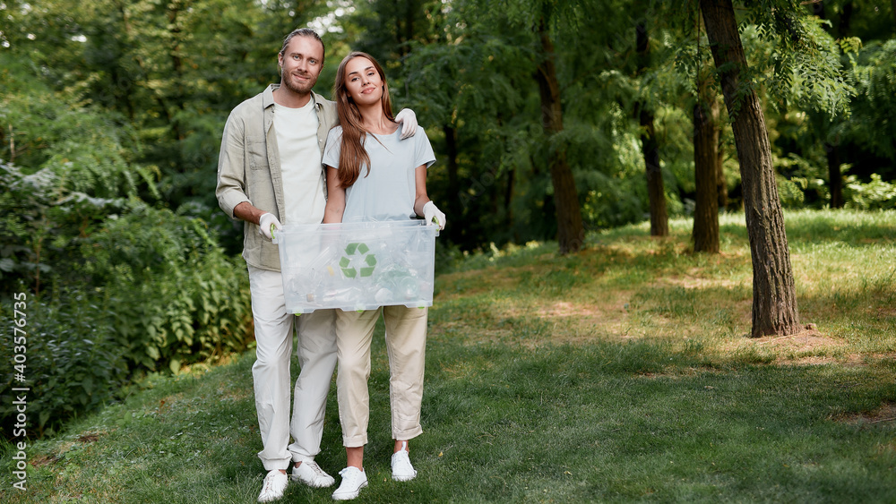 Save the planet from plastic. Young happy couple, eco volunteers holding recycle bin and smiling at camera while cleaning together green forest from plastic waste Save the planet from plastic. Young happy couple, eco volunteers holding recycle bin and smiling at camera while cleaning together green forest from plastic waste