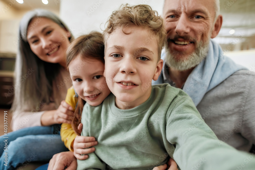 Happy family, grandparents with two cute kids making a selfie on smartphone while having fun in the living room, using modern technologies