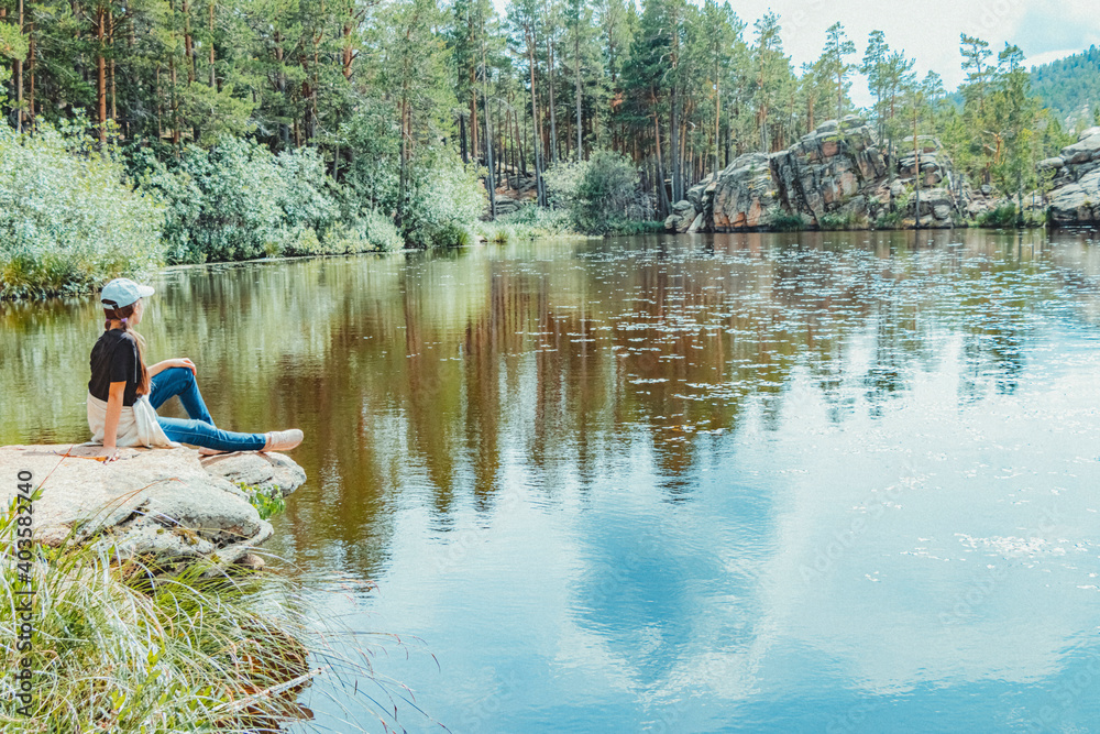 Young beautiful girl in jeans and baseball cap sits on stone on shore of lake surrounded by forest and rocks. Young beautiful girl in jeans and baseball cap sits on stone on shore of lake surrounded by forest and rocks.