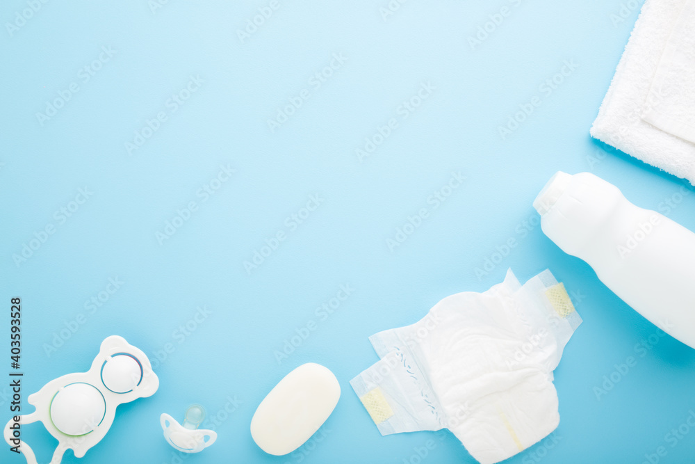 White bottle of talcum powder, diaper, towel, soap, rattle and soother on light blue table background. Pastel color. Products of baby daily hygiene. Closeup. Empty place for text. Top down view.