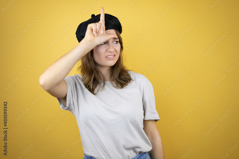 Beautiful young caucasian girl wearing french look with beret over isolated yellow background making fun of people with fingers on forehead doing loser gesture mocking and insulting.