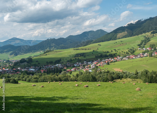 view on village Liptovska Luzna at the foothills of Low Tatras mountains with lush green meadow, forested hills and straw bales. Slovakia, summer sunny day, blue sky white clouds background