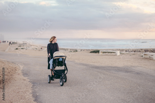 A young woman in warm dress with a baby carriage walking along a beach
