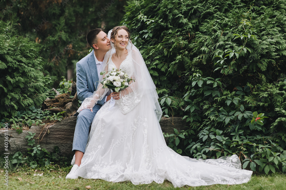 A young groom in a blue striped suit and a beautiful curly-haired bride in a lacy white dress are sitting on a log, against the background of green bushes in nature. Wedding portrait of the newlyweds.