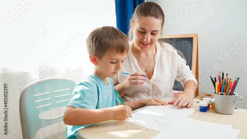 Smiling mother looking at her little son drawing or writing while sitting behind desk at home classroom. Concept parents and children of education and remote school at home during lockdown and self