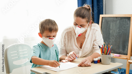 Young female teacher and little boy wearing protective mask respirators studying at school classroom. Parents protecting children from virus during lockdown and self isolation. Remote school and