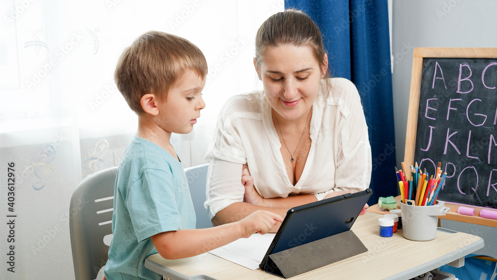 Little boy studying on digital tablet computer at home. Mother looking at er little son doing homework on tablet computer Little boy studying on digital tablet computer at home. Mother looking at er little son doing homework on tablet computer