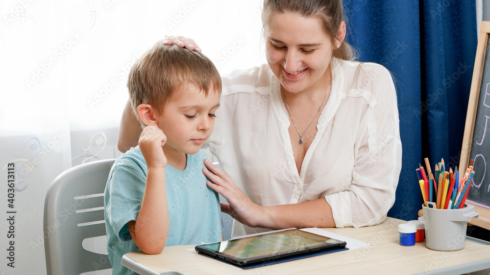 Smiling mother hugging and caressing her smart child studying on tablet computer. Little boy doing homework on digital tablet computer at home