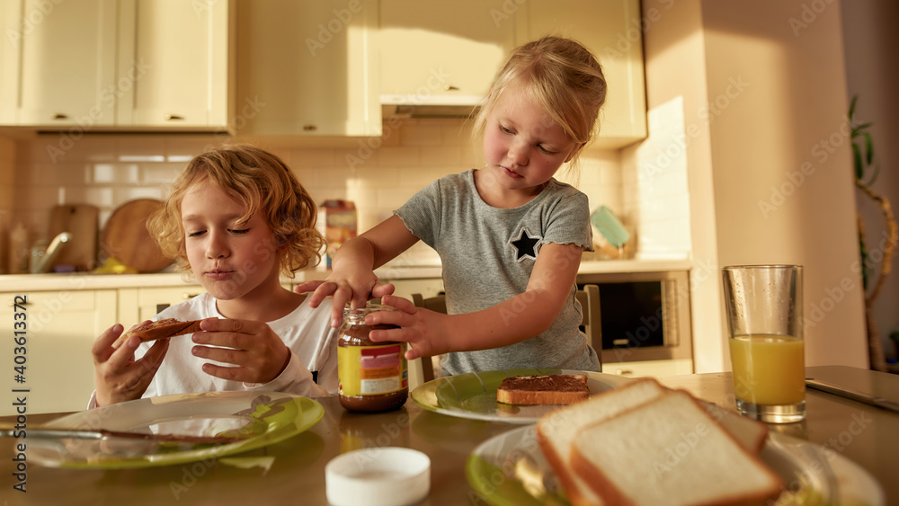 Two adorable little kids spreading chocolate butter on toasted bread while preparing breakfast for themselves in the kitchen