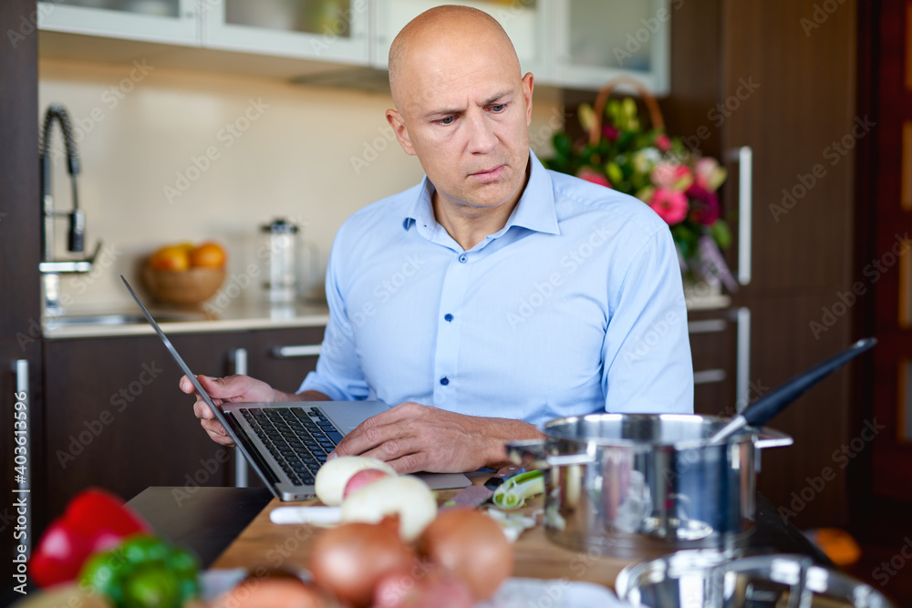 Brutal man in kitchen preparing food and uses laptop. Brutal man in kitchen preparing food and uses laptop.