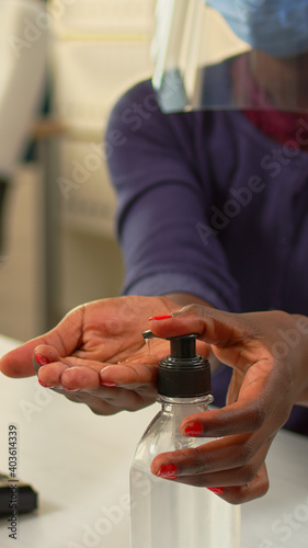 Close up of black woman using hand sanitizer while working in office room in new normal company workplace. African employee cleaning disinfecting hands using alcohol gel against corona virus.