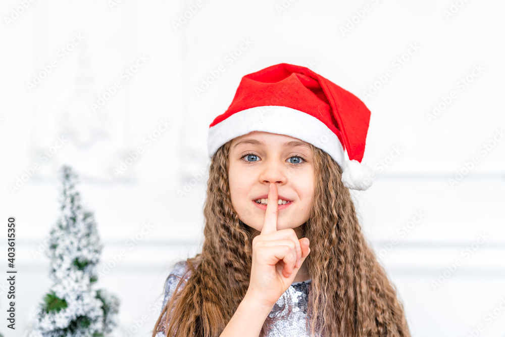Charming curly haired little girl wearing a Santa hat smiles and looks at the camera in a bright studio with Christmas decorations Charming curly haired little girl wearing a Santa hat smiles and looks at the camera in a bright studio with Christmas decorations