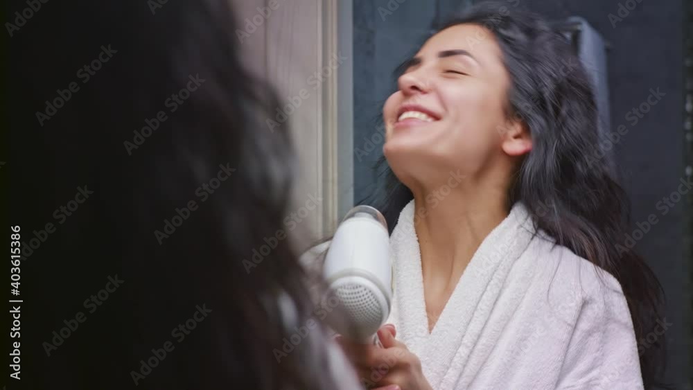 Beautiful caucasian woman looking at her reflection in the mirror and singing while drying hair with blow dryer after taking shower in bathroom Beautiful caucasian woman looking at her reflection in the mirror and singing while drying hair with blow dryer after taking shower in bathroom