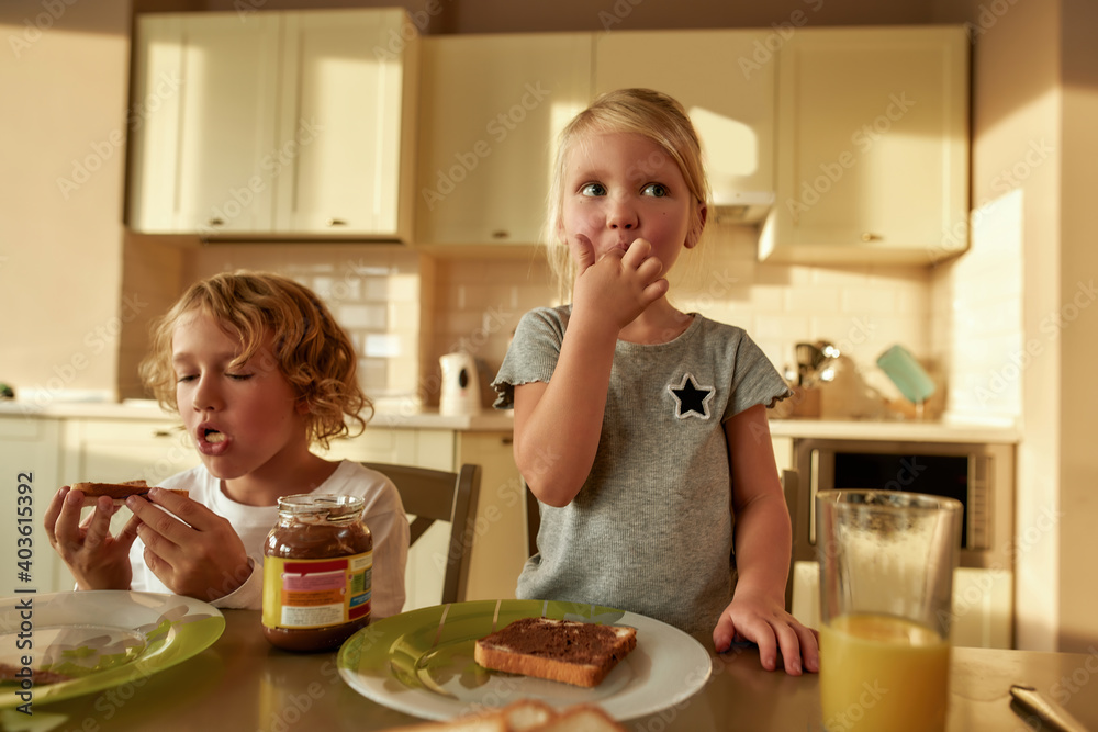 Two adorable little kids eating toasted bread with chocolate butter while having breakfast in the kitchen