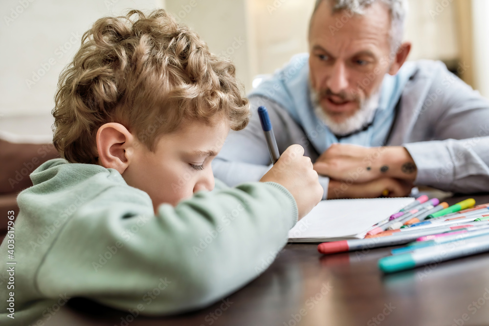 Grandad with serious face helping his little grandson drawing with colored pencils, spending time at home Grandad with serious face helping his little grandson drawing with colored pencils, spending time at home