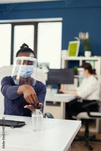 African woman in corporation office applying hand sanitizer wearing face mask during coronavirus. Businesswoman in new normal workplace disinfecting while colleagues working in background.