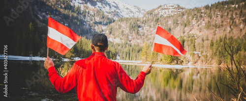 male hiker is holding two austrian flags and is standing in front of mountain lake somewhere in the alps photo