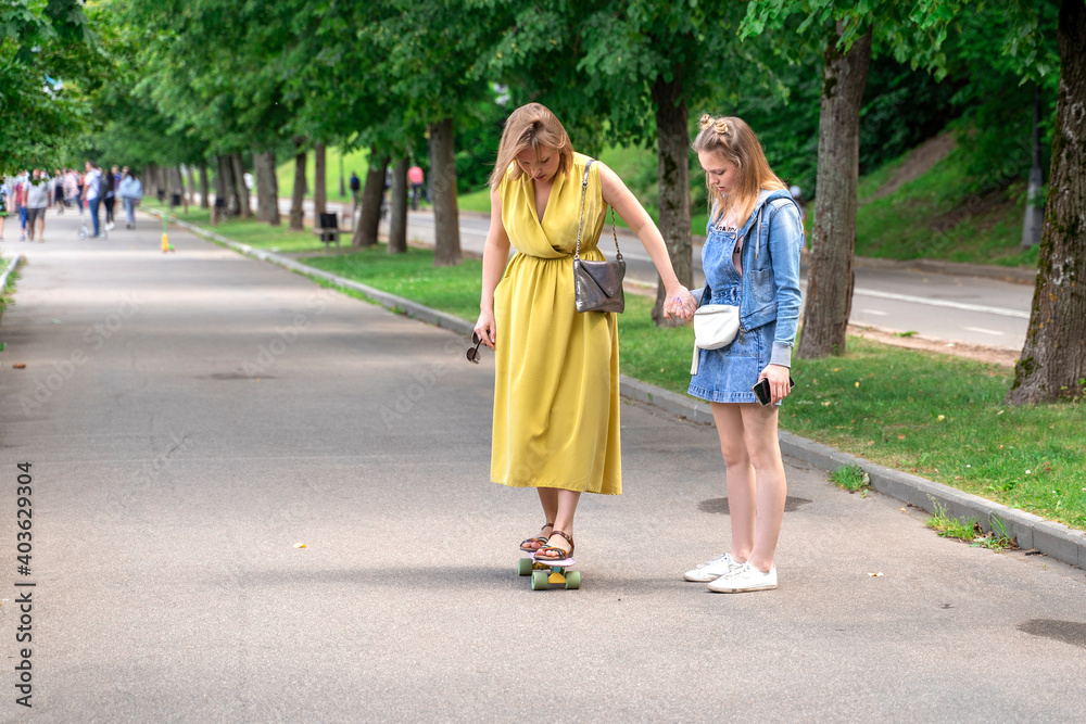 woman standing on longboard supported by her daughter. Woman learning to skateboard outdoors. woman standing on longboard supported by her daughter. Woman learning to skateboard outdoors.