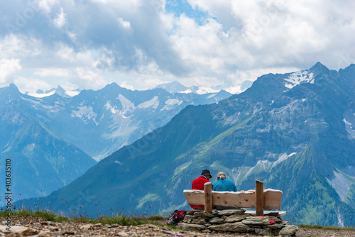 Two older people, tourists, rest on a bench in the mountains. Around them there is a view of the majestic snow-covered Alps.