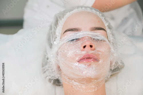 Treatment, cure of face skin in cosmetology clinic. Beauty procedure with problem skin for young woman. Woman with cream with anesthesia and plastic film on her face before PRP therapy procedure.