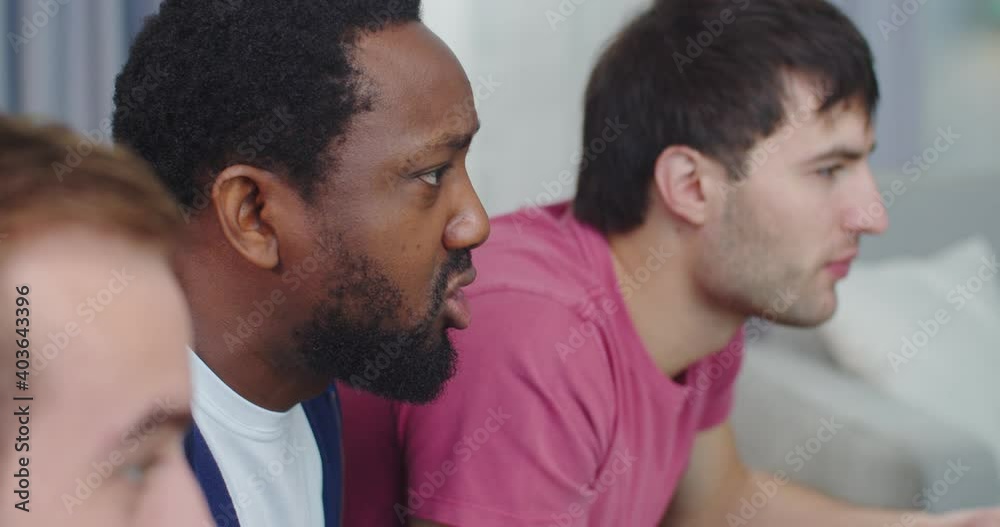 Side view. Close up of African American and Caucasian guys best friends at home watching football game on TV together and screaming, cheering for soccer team on sport channel. Fans concept Side view. Close up of African American and Caucasian guys best friends at home watching football game on TV together and screaming, cheering for soccer team on sport channel. Fans concept