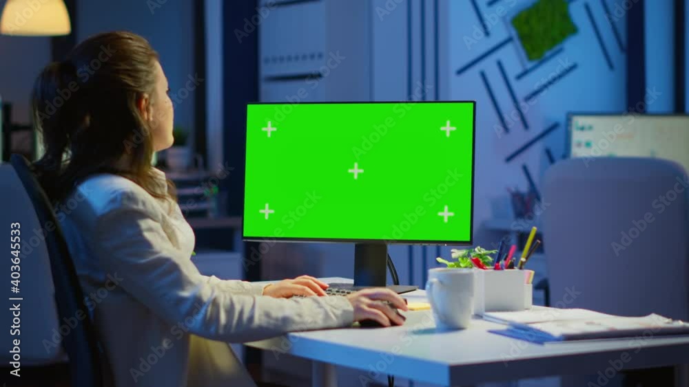 Businesswoman looking at green screen monitor of computer sitting at desk in business office late at night. Freelancer watching desktop monitor display with green mockup, chroma key working overtime.