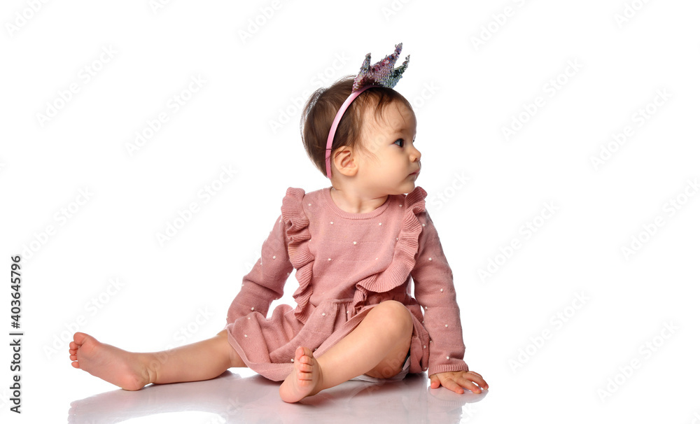 little cute baby in a headband in the form of a crown sits in a pink dress on a gray background, looks to the side at an empty seat. dad and mom princess little cute baby in a headband in the form of a crown sits in a pink dress on a gray background, looks to the side at an empty seat. dad and mom princess