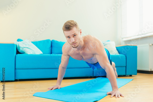 A muscular athletic man in shorts is energetically starting to do fitness exercises at home in his apartment with a modern interior.