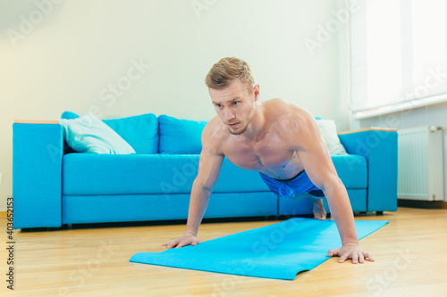 A muscular athletic man in shorts is energetically starting to do fitness exercises at home in his apartment with a modern interior.