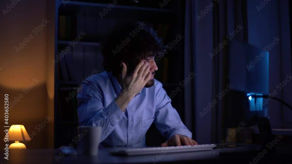 Sleepy young bearded man working late at night on computer rubbing eyes at home office in dark room. Tired sleepy student prepares for late night session. Young freelancer working late over project. Sleepy young bearded man working late at night on computer rubbing eyes at home office in dark room. Tired sleepy student prepares for late night session. Young freelancer working late over project.