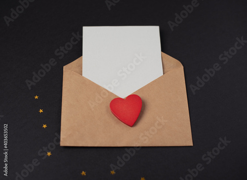 Craft envelope with a blank sheet of paper inside and red wooden heart on the black background. Romantic love letter for the Valentine's day concept. Space for text.