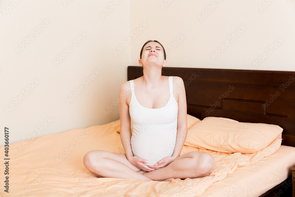 Young pretty pregnant woman, in pain, sitting on bed in bedroom, preparing to give a birth Young pretty pregnant woman, in pain, sitting on bed in bedroom, preparing to give a birth