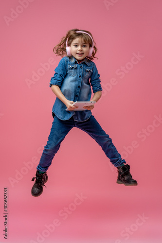 little girl in headphones and with tablet computer, jumping on pink background