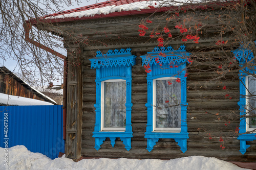 windows of a wooden house with blue carved platbands on a winter day. rowan with red berries grows under the windows. Cozy home © olgaS