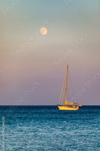 Full moon rising over the water with a small sailing boat in the foreground. Sailing boat with raising moon at sunset. Moon rising over the sea and yacht floating on the water surface. Sardinia, Italy