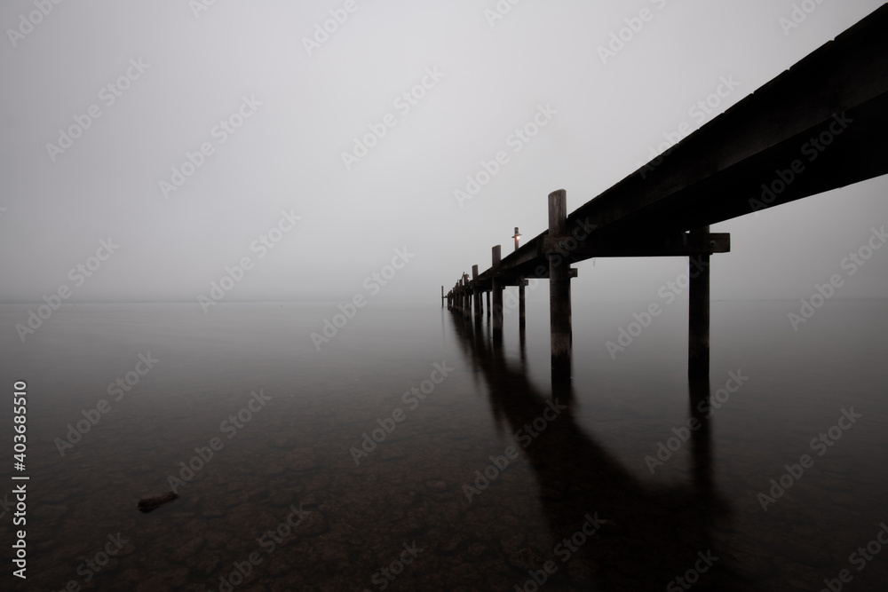 Mole (pier) on the „Chiemsee“ lake in Bavaria, Germany. Wooden bridge in winter time with frozen and icy lake and dim lanterns for fishing. Dark and Foggy lake.

