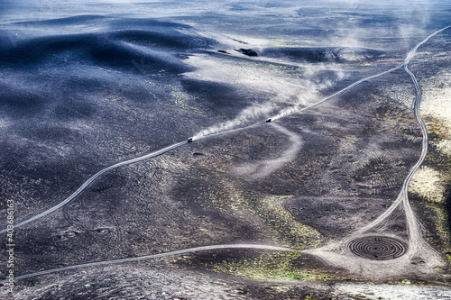 black slag cone in the Tolbachik valley. bright spots of moss.
two cars are driving in clouds of dust.