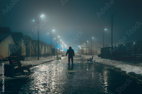 Silhouette of dark man in hood in night illuminated city alley in foggy weather, misty horror and scary atmosphere concept.