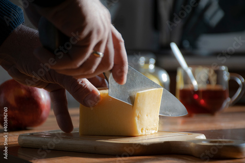 A man's hand with a knife cuts the cheese in focus. Mug of tea, lemon and apple in the background out of focus.