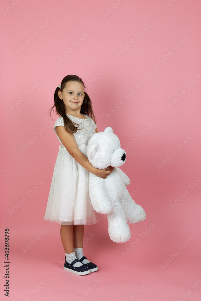 A full-length mischievous, beautiful girl with ponytails and a white princess dress holds a white teddy bear isolated on a pink background. A full-length mischievous, beautiful girl with ponytails and a white princess dress holds a white teddy bear isolated on a pink background.
