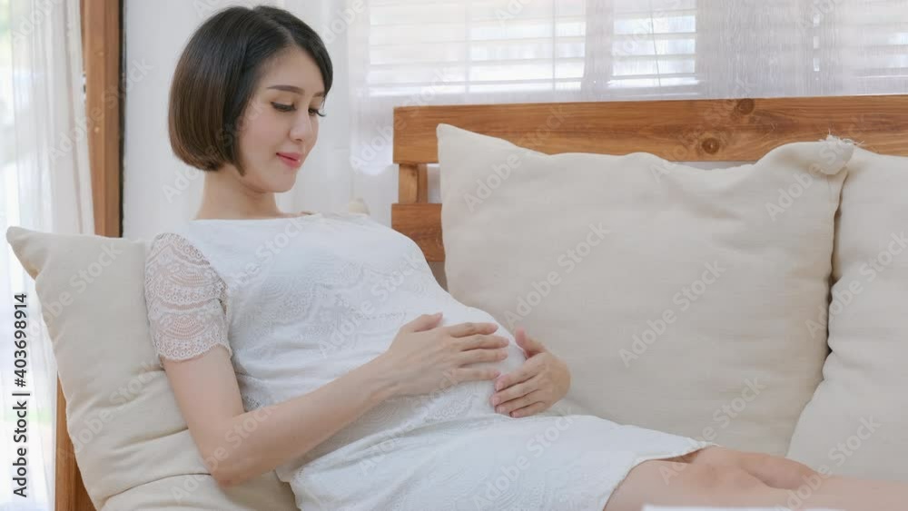 Asian beautiful pregnant woman sit on sofa and touch belly in living room and she express happiness emotion during stay at home. Healthcare support pregnancy family concept Asian beautiful pregnant woman sit on sofa and touch belly in living room and she express happiness emotion during stay at home. Healthcare support pregnancy family concept