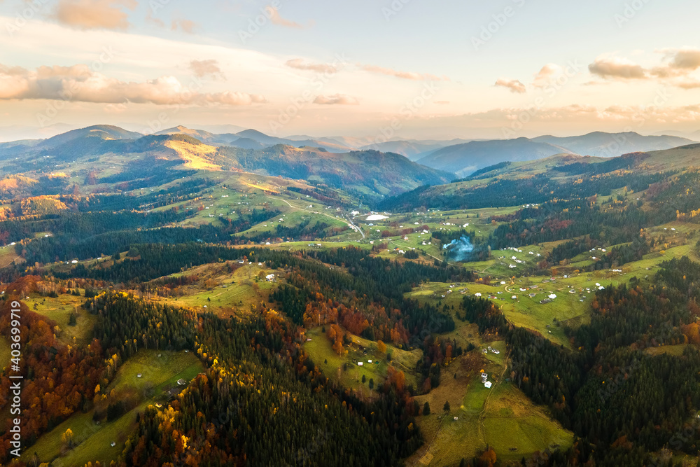 Aerial view of distant village with small shepherd houses on wide hill meadows between autumn forest trees in Ukrainian Carpathian mountains at sunset.