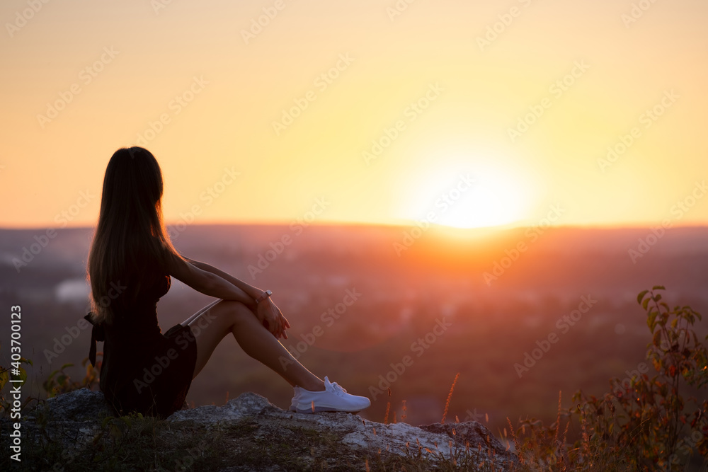 Young slim woman in black short dress sitting on a rock relaxing outdoors at summer sunset. Fashionable female enjoying warm evening in nature.