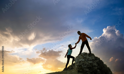 Φωτογραφία Man and woman hikers helping each other to climb a big stone at sunset in mountains