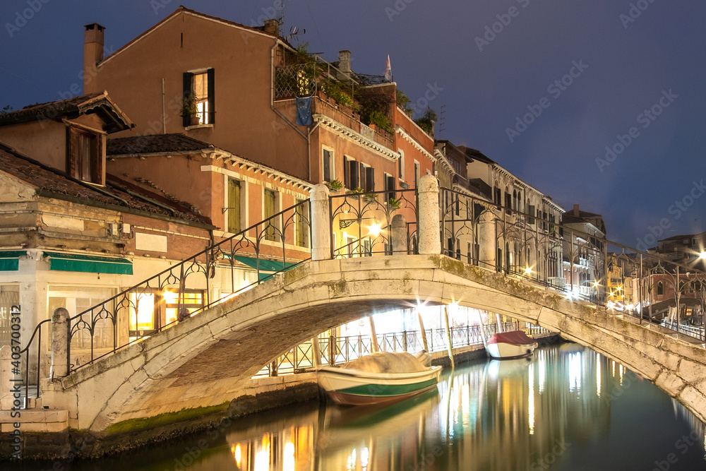 Evening view of illuminated old buildings, bridges, floating boats and light reflections in canals water in Venice, Italy.
