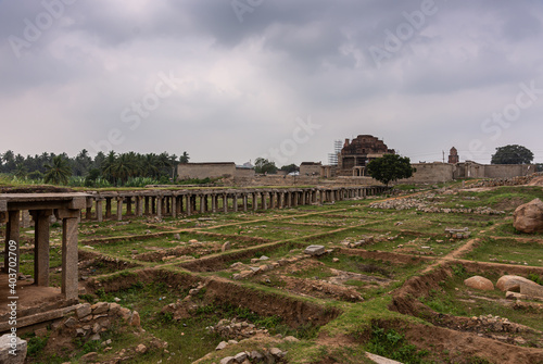 Hampi, Karnataka, India - November 5, 2013: Sri Krishna tank in ruins. Old fundaments form squares in grass. Gallery and entrance damaged gopura to temple proper under gray cloudscape. photo