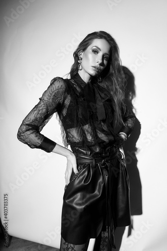 Elegant woman in black lace blouse, tights, blazer, leather shorts and sabot shoes standing and posing against white background. Hard light