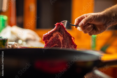 Man is seen lifting a hefty juicy beef steak with a fork and putting it into a pan in a kitchen